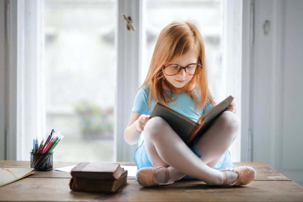 A young girl wearing glasses with hyperlexia reading a book in her room