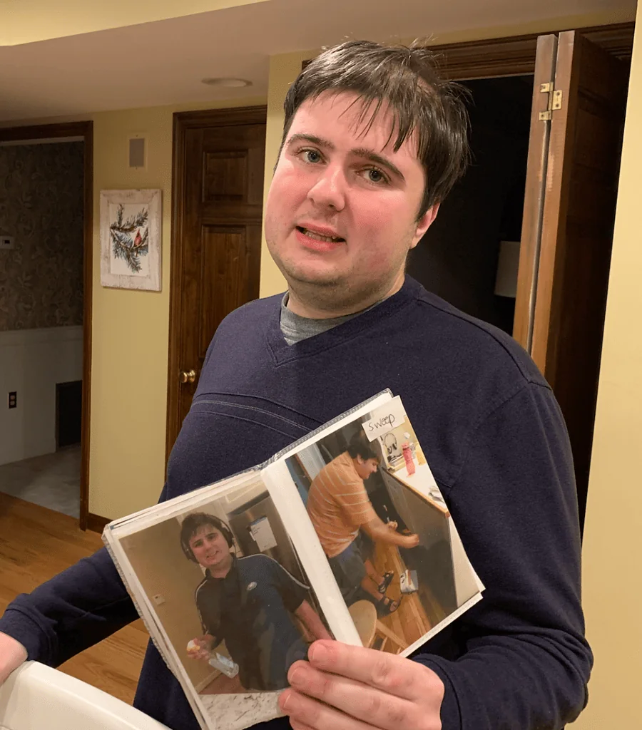 Lucas Barbera holding up his morning routine activity schedule book to show two steps after eating breakfast which include wiping the counter and sweeping the floor.