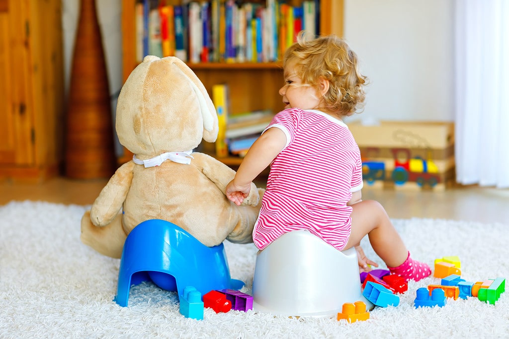 Potty training and constipation photo of girl on toilet, smiling to teddy bear